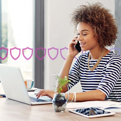Photo of a woman in an office setting talking on her phone while typing or scrolling on her laptop. In the background, a series of small purple shields runs across the frame, representing security.