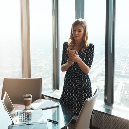 Photo of woman in an office. She is looking at something on her cellphone. On a table in the foreground are her laptop and some papers.