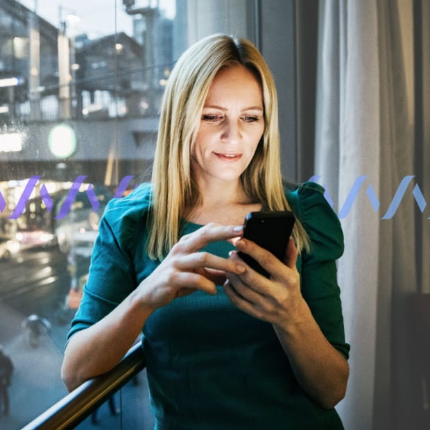 Photo of a woman standing by a window in a hotel room scrolling on her cell phone.